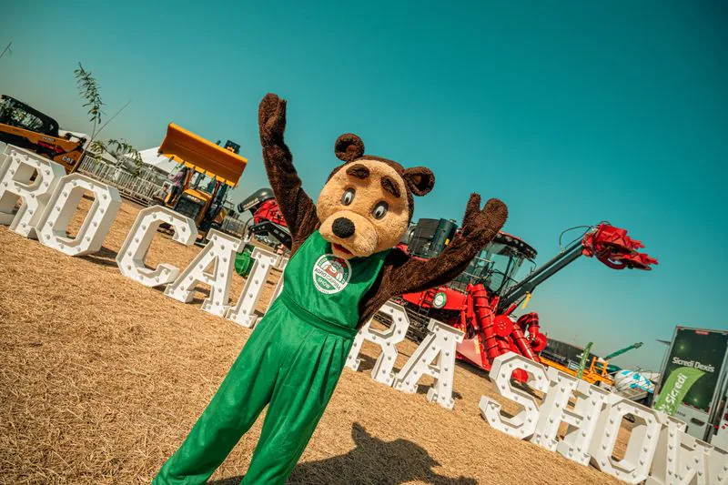 Mascote da feira celebrando entre máquinas e visitantes, edição 2024